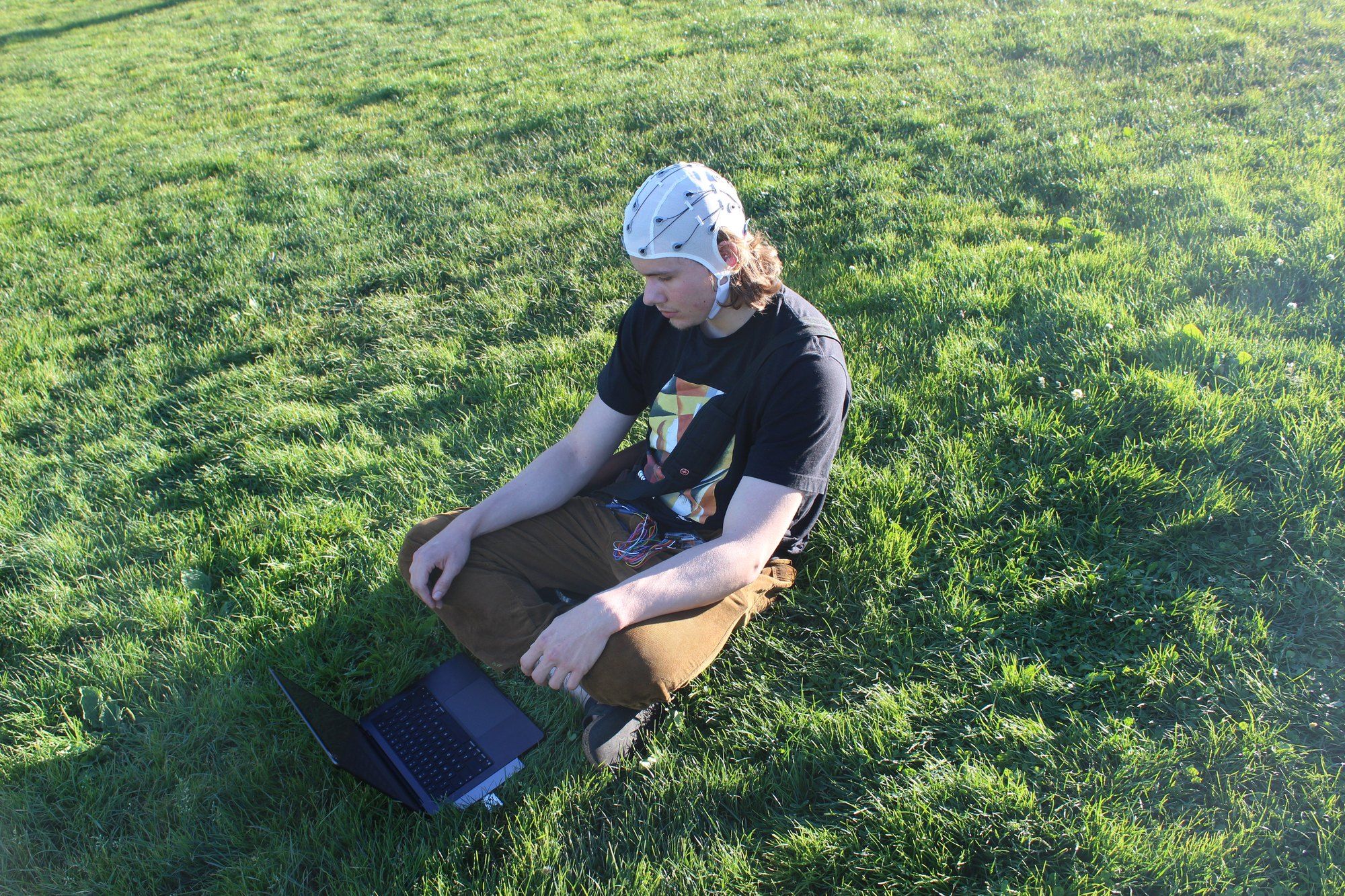 Chris sits cross-legged on the grass in Dolores Park in a white EEG research cap, its wires running to a laptop on the grass in front of him